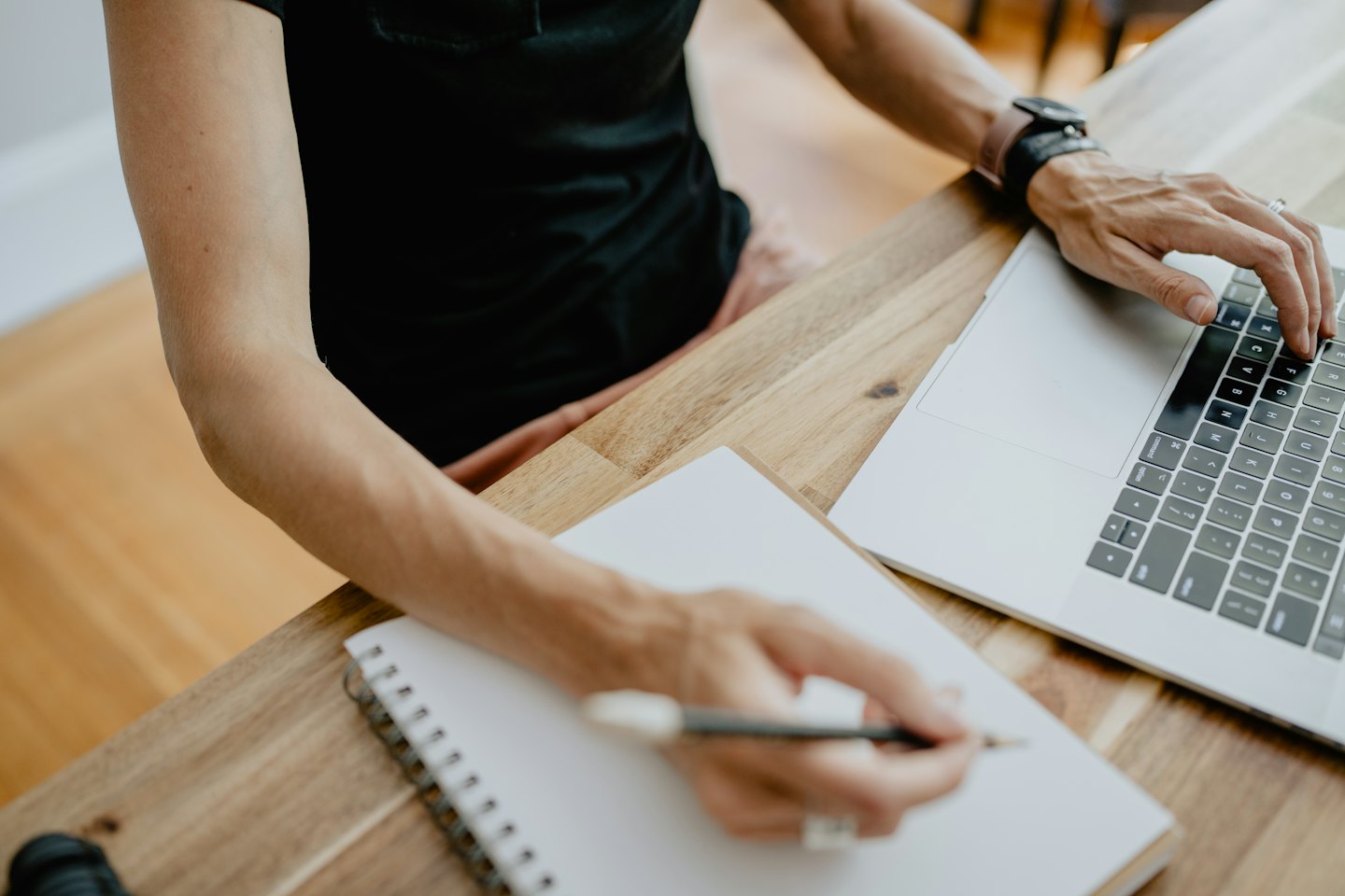 Tangan mengetik di laptop dan menulis di buku catatan.